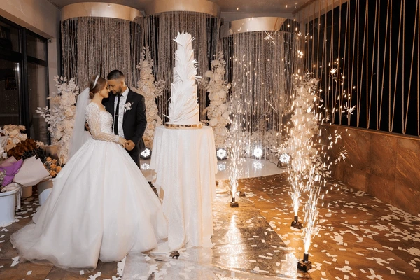 Bride and groom standing beside their wedding cake while indoor spark fountains illuminate the room